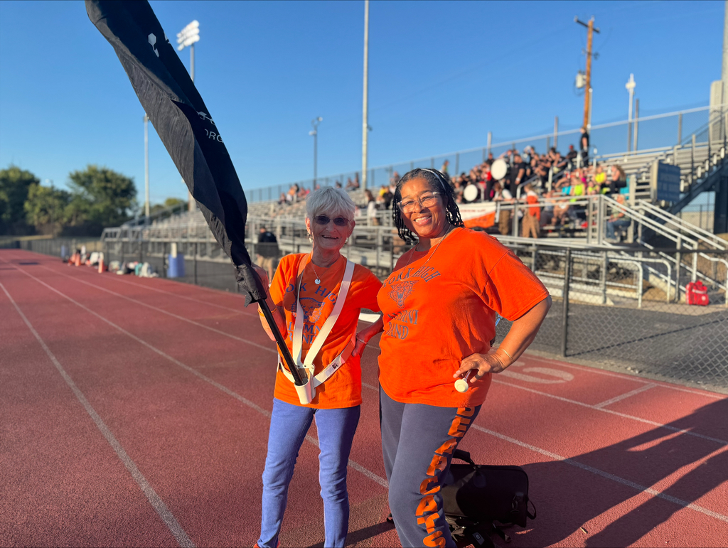 Two individuals in orange shirts stand on a track, one holding a black flag, with an audience and band visible in the background.