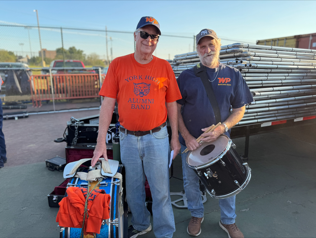 Two York High Alumni band members standing beside stacked equipment, one holding a drum and the other wearing an orange alumni band t-shirt.