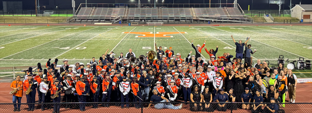 A large group of York City School District musicians in colorful uniforms gathering on a football field, posing joyfully with instruments under stadium lights.