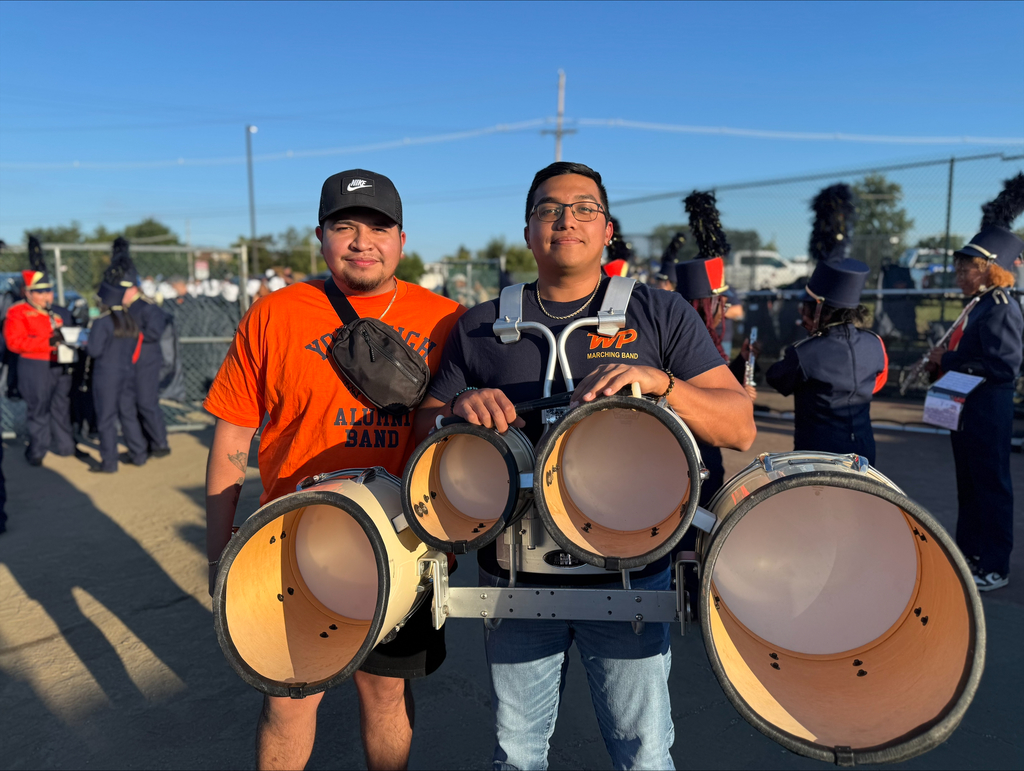 A York High Alumni band member and a band student posing together and holding instruments in an outdoor setting. People can be seen in the background.