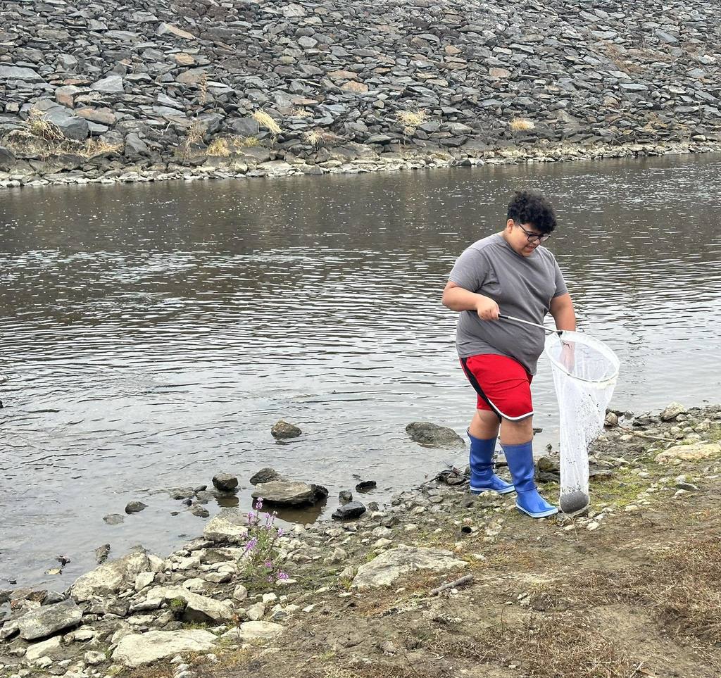 A student holding a white net filled with materials near a lake surrounded by large rocks.