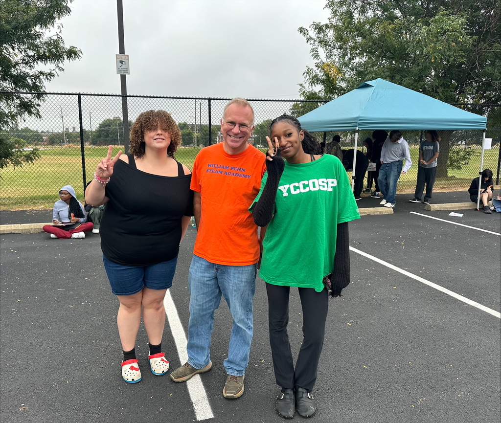 Two students and a district staff member posing together in an outdoor setting. Both of the students are holding up the peace sign. Other people can be seen in the background.