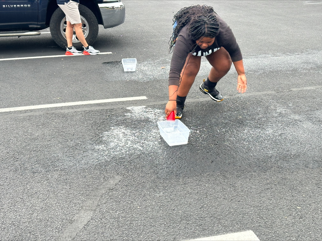 A student dumping a cup of water into a bucket in an outdoor setting. Another person can be seen in the background.