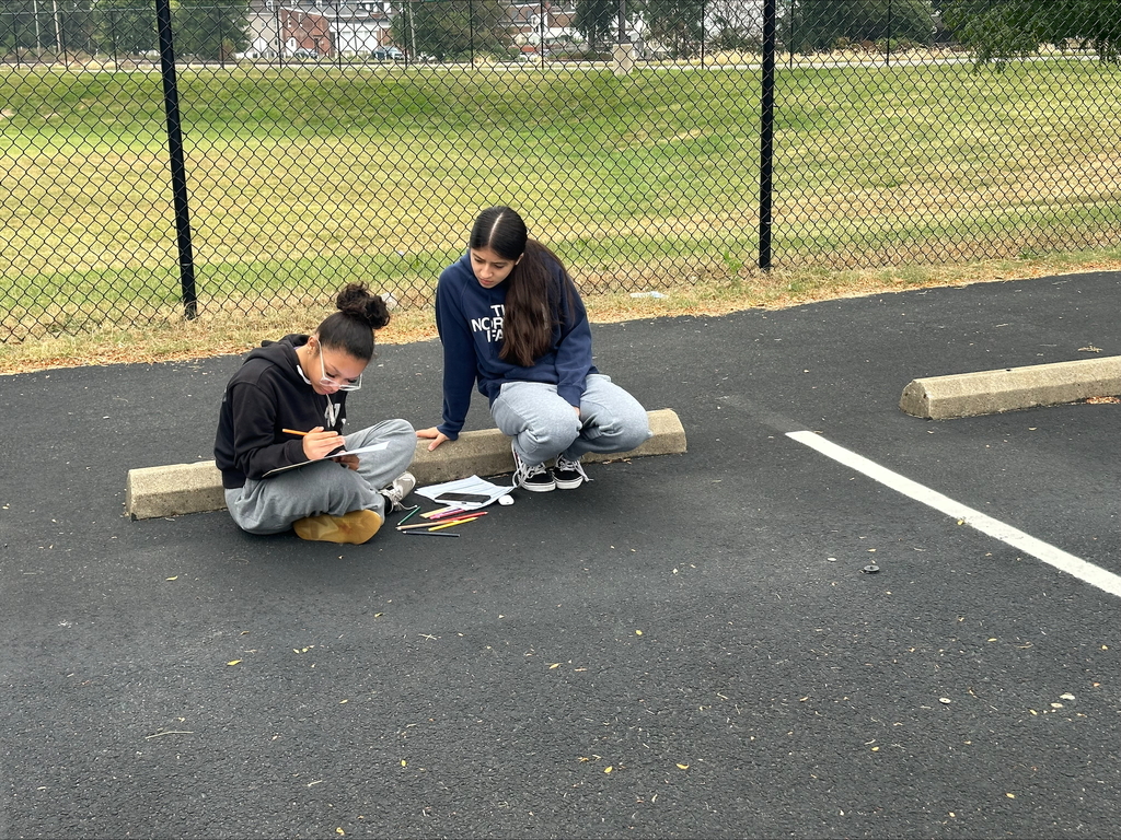 Two students sitting in an outdoor setting working on an assignment together. One of the students is writing on a piece of paper.