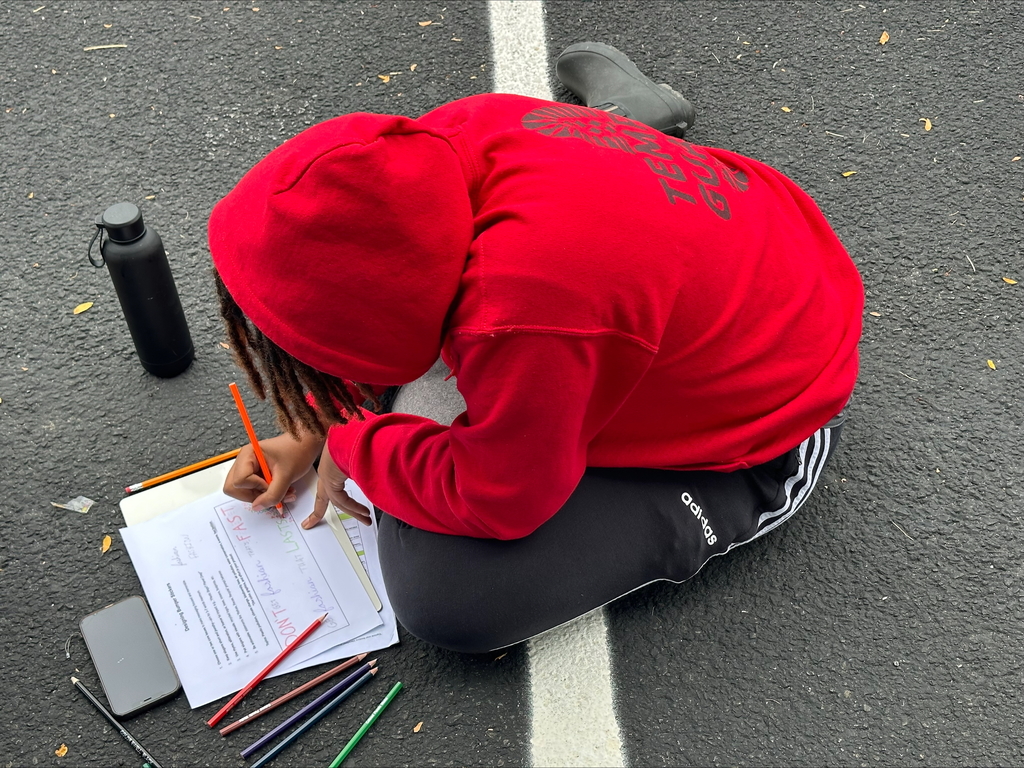 A student sitting in an outdoor setting writing on a piece of paper with colored pencils. A water bottle and a phone is nearby the student.