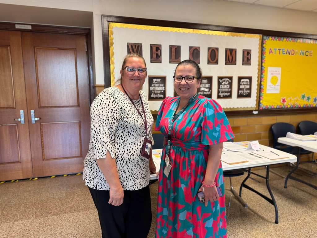 Davis PreK-8 Principal, Ms. Sweitzer and a district staff member standing in a hallway with a welcome sign on the wall. Both women are near tables that have papers on them.
