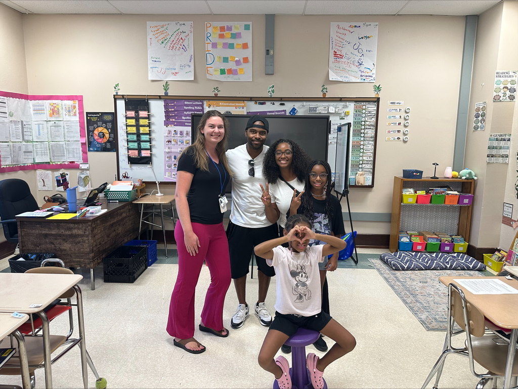 Three adults and two children are posing for a group photo in a classroom setting. One of the children is forming a heart with her hands.