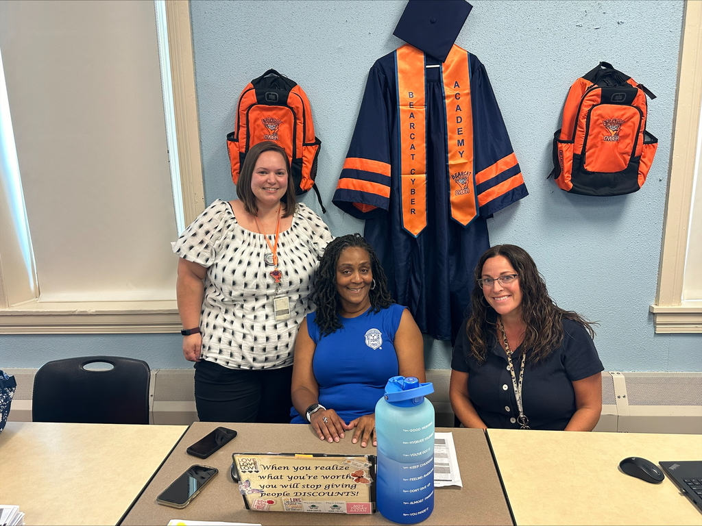 Three district staff members posing behind a table with an inspiring sign and large water on it. Two of the staff members are sitting down and one is standing up. Bearcat Cyber Academy's robe and two backpacks can be seen hanging on the wall behind them.