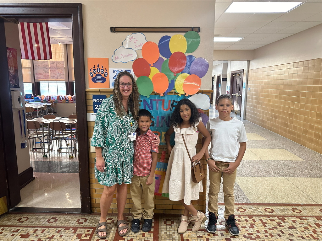 A district staff member and three children are posing in a school hallway in front of a wall that displays colorful balloons and a sign with positive messages.