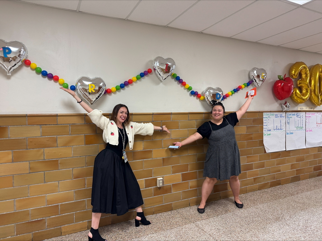Two district staff members are enthusiastically posing in a decorated hallway with their hands lifted up in the air.