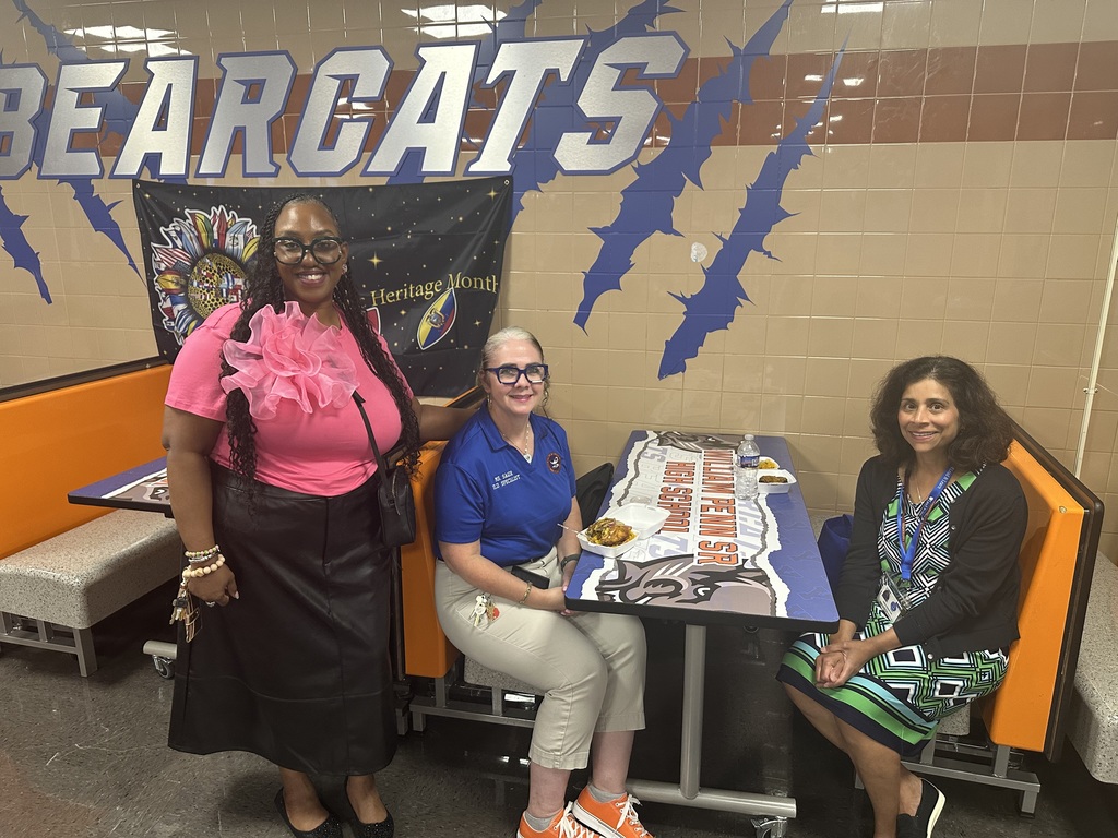 William Penn Senior High School's 9th grade Principal, Mrs. Floyd standing next to two district staff members who are sitting down at a table with food on it. The women are standing in front of a wall labeled "Bearcats".