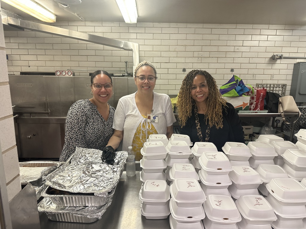 Three district staff members standing in a kitchen area, surrounded by stacked food containers and pans that are covered with foil.