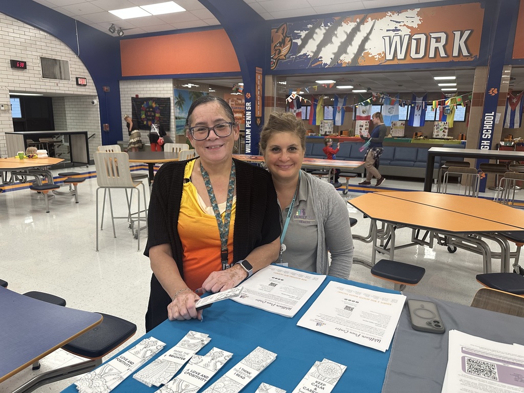 District staff members standing at a table with papers on it in a school cafeteria. The background features flags with a "WORK" sign on it.