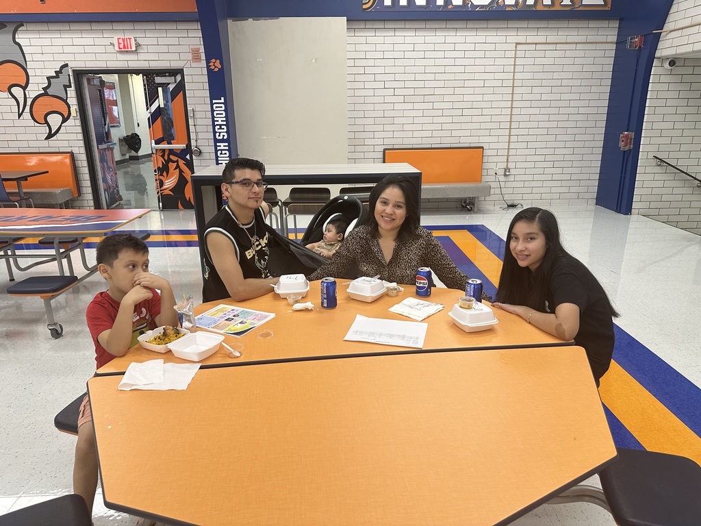 A family of five, including a baby, is sitting at an orange table in a school cafeteria enjoying food and drinks.