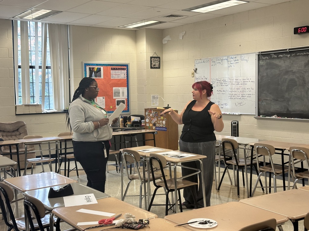 Two people are having a conversation in a classroom surrounded by desks and chairs. One of the people is holding papers, and the other person is using hand gestures.