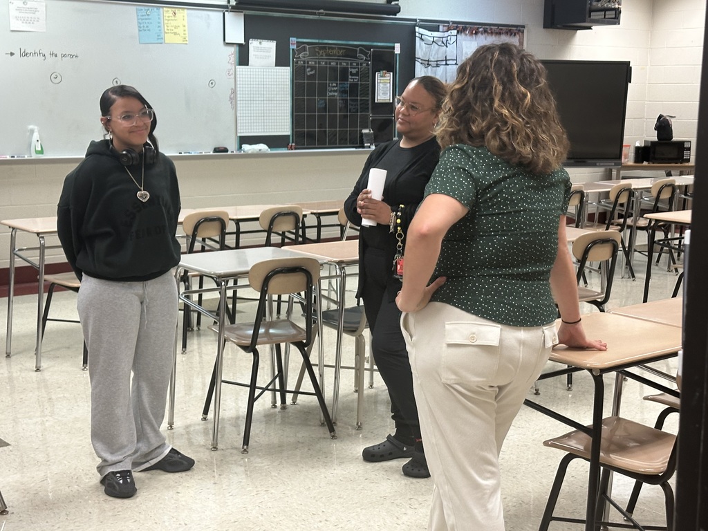 Three people standing and having a conversation in a classroom surrounded by desks and chairs.