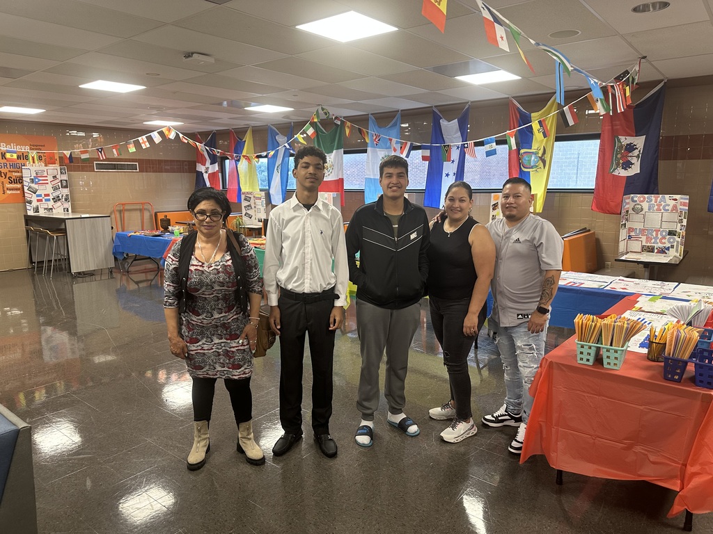 A group of five people are standing in a decorated area with international flags hanging above. There are tables displaying colorful items and decorations.