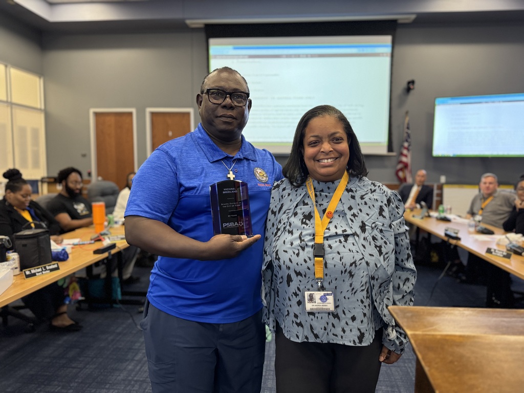 Dr. Andrea Berry-Brown, Superintendent of Schools standing next to Mr. Michael Breeland, School Board Director in a conference room. Director Breeland is holding a plaque in his hand. Other people are seated at tables in the background.