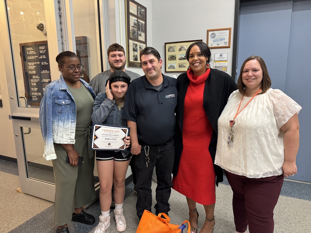 District leadership standing next to two adults and one student in a lobby area. The student is holding up a certificate that is titled "Certificate of Appreciation."