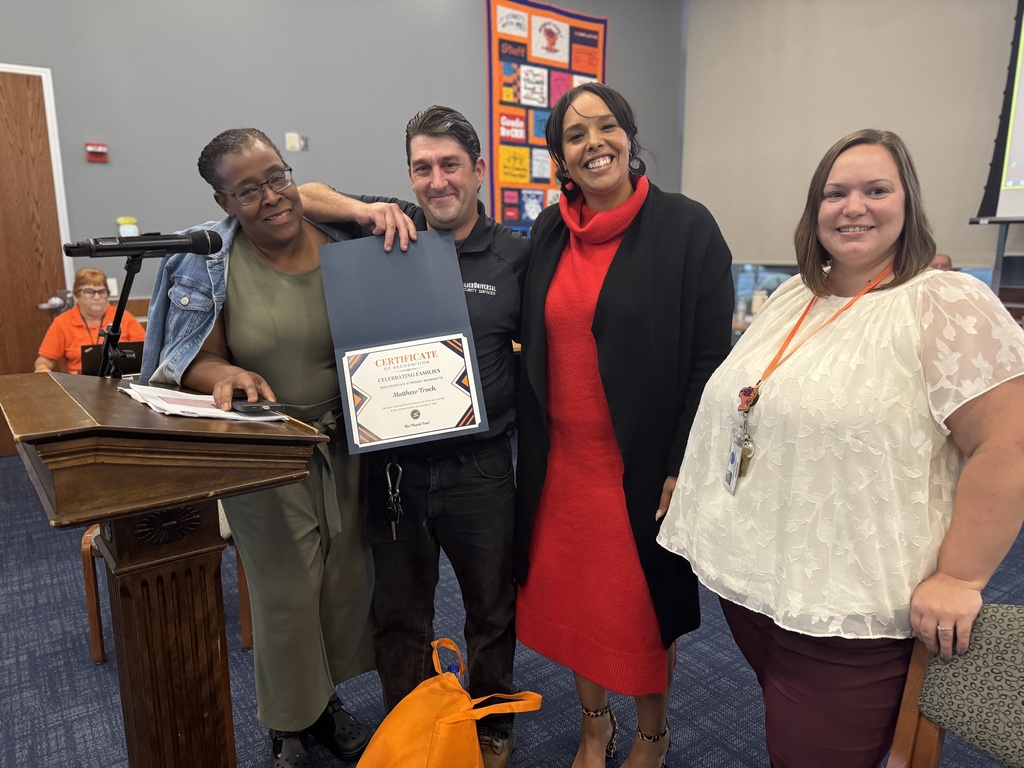 District leadership standing with a district parent who is holding a certificate in his hand next to a podium in a conference room. The certificate is titled "Certificate of Appreciation." A person can be seen in the background.