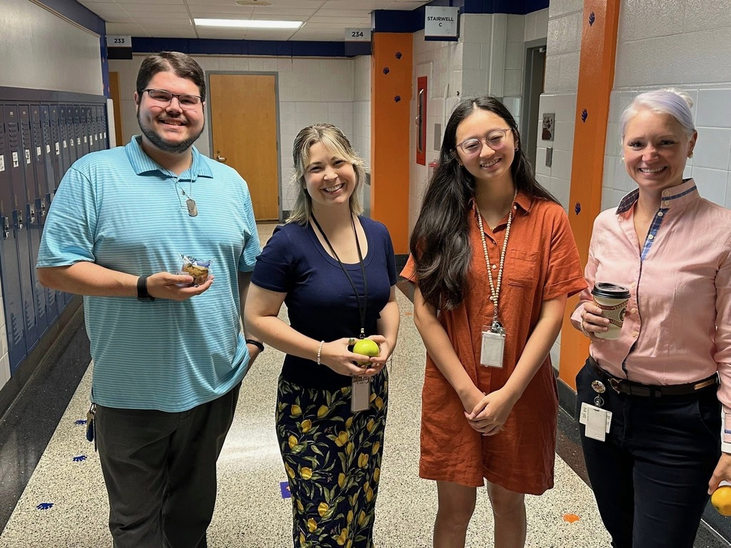 Four district staff members are smiling while standing in a school hallway near lockers. One person is holding a muffin, another person is holding an apple, and a third person is holding a cup.