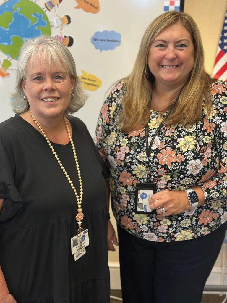 Two district staff members are smiling while standing in front of a wall that has a globe on it and cartoon children on it.