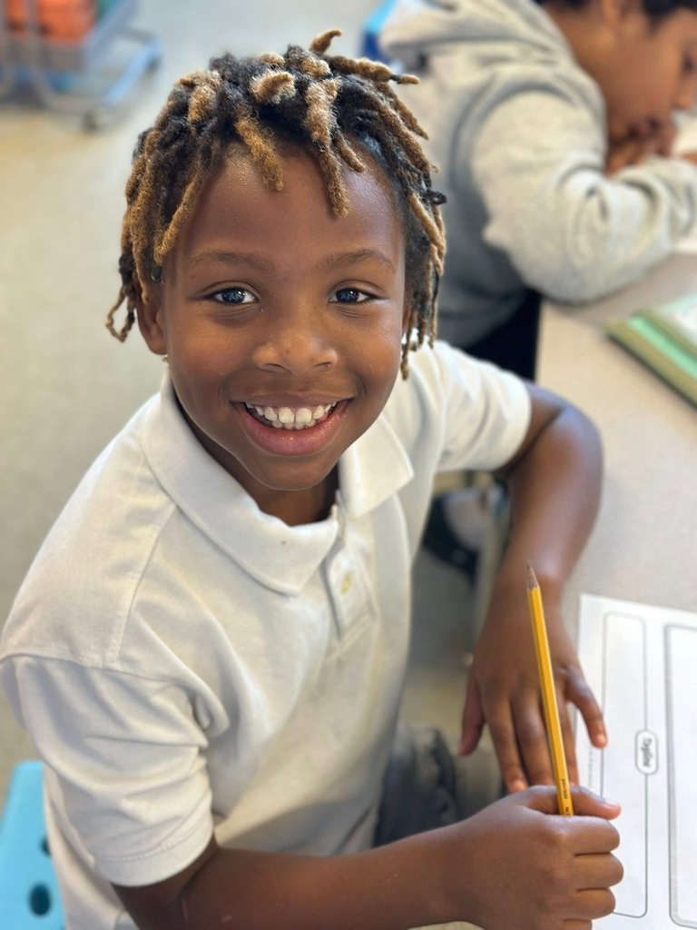 A student smiling and holding a pencil in his hand in a classroom setting. Another student can be seen in the background.