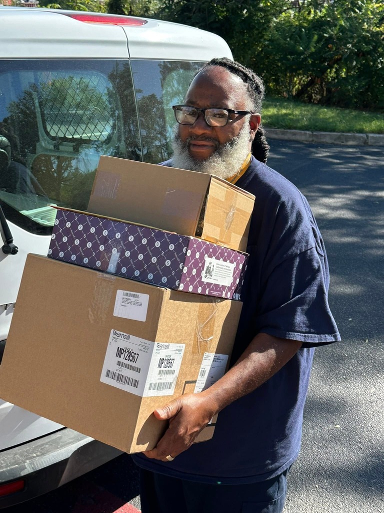 A district staff member is carrying three cardboard boxes while standing behind a white van outside.