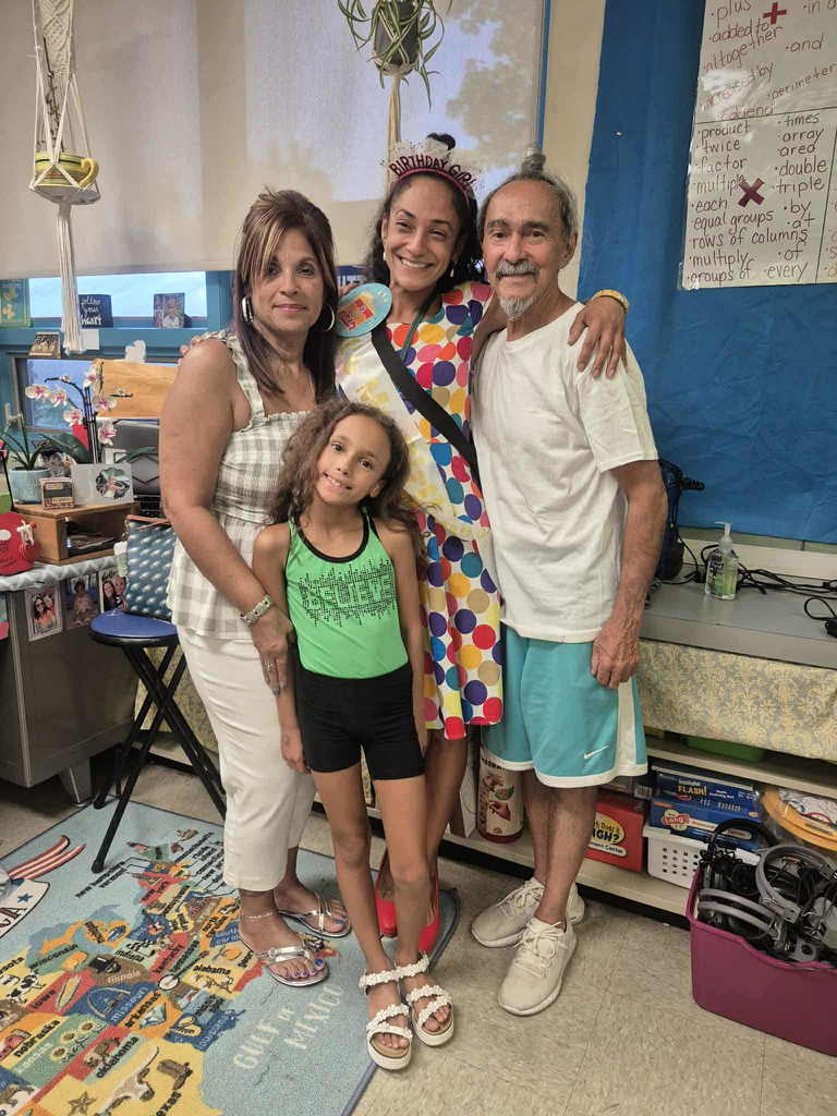 Three adults and one child are smiling in a classroom setting while standing on a display of a map. One of the woman is wearing a birthday crown and a birthday sash.