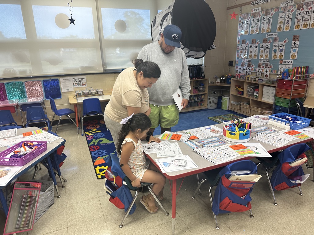A young student is sitting at a classroom table filled with colorful worksheets and supplies. Two adults are standing by her looking down at the table.