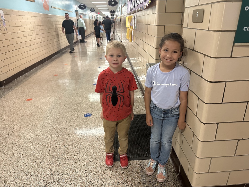 Two students are smiling in a school hallway next to a wall. Other people can be seen in the background.