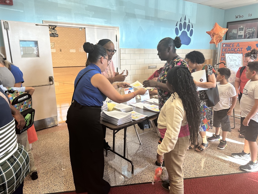 A school event with district leadership distributing papers to a woman at a table. Other people can be seen in the background.