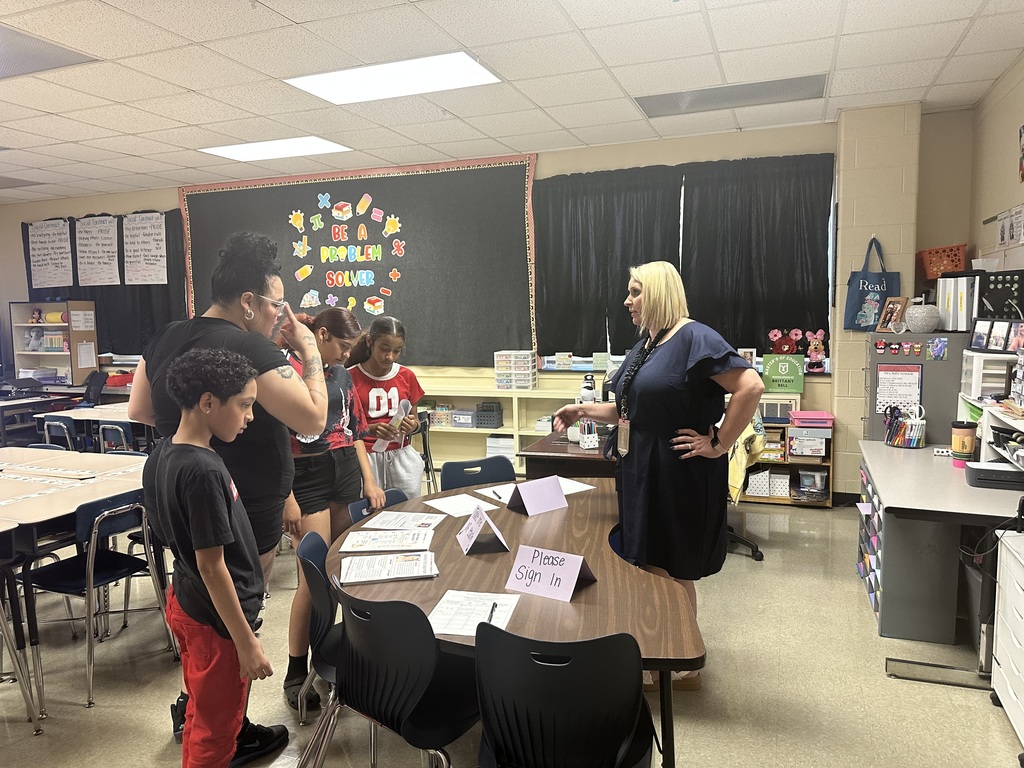 A district staff member is having a conversation with a woman while three children are standing nearby in a classroom.
