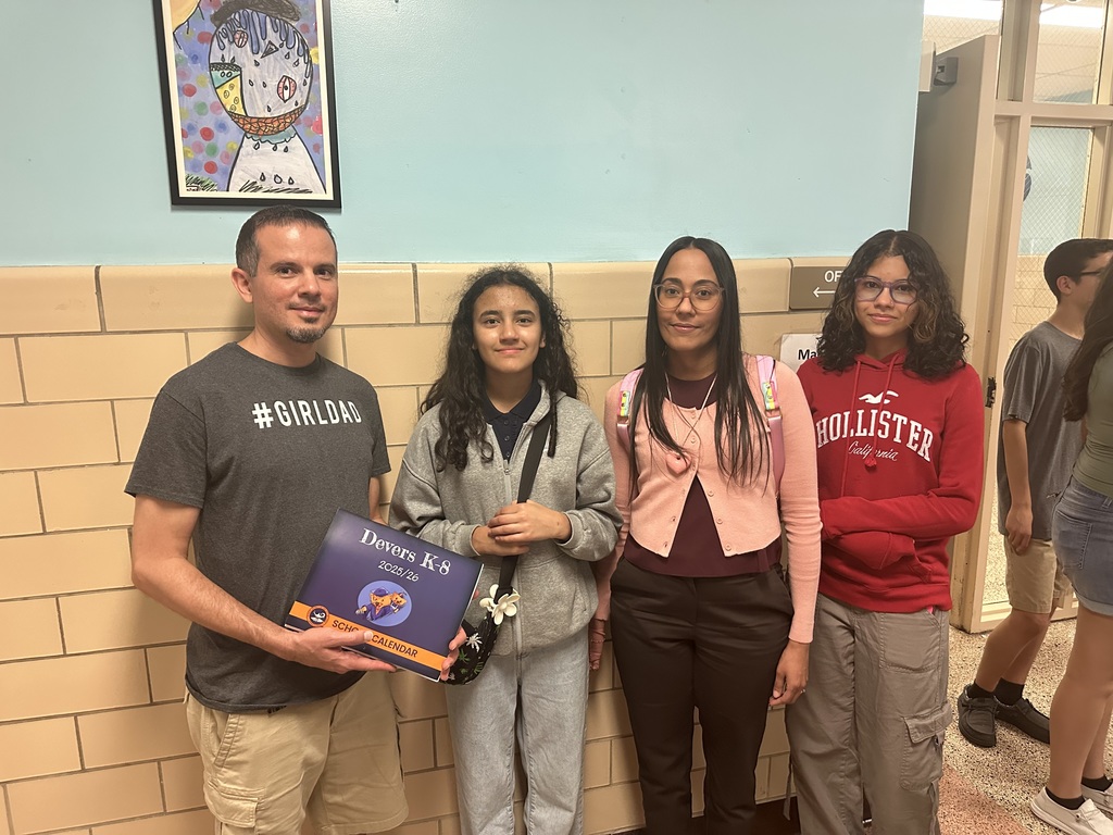 A family of four, two adults and two students are standing in front of a wall in a school hallway. A picture can be seen above the man.