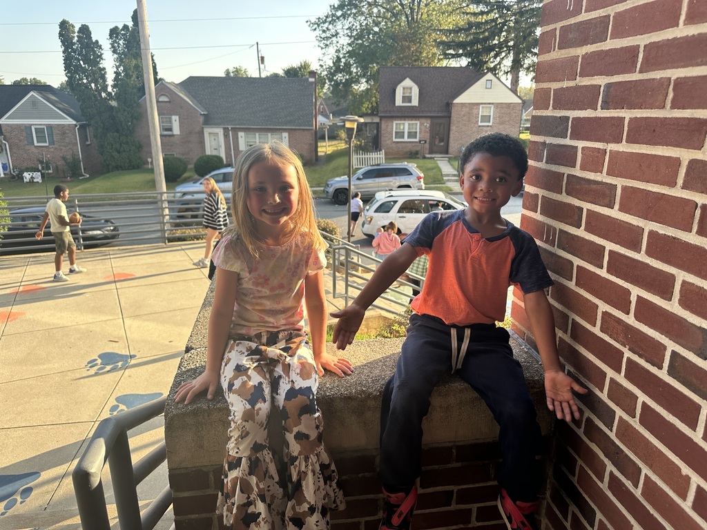 Two young students are sitting on a brick ledge outside. Houses and other people can be seen in the background.