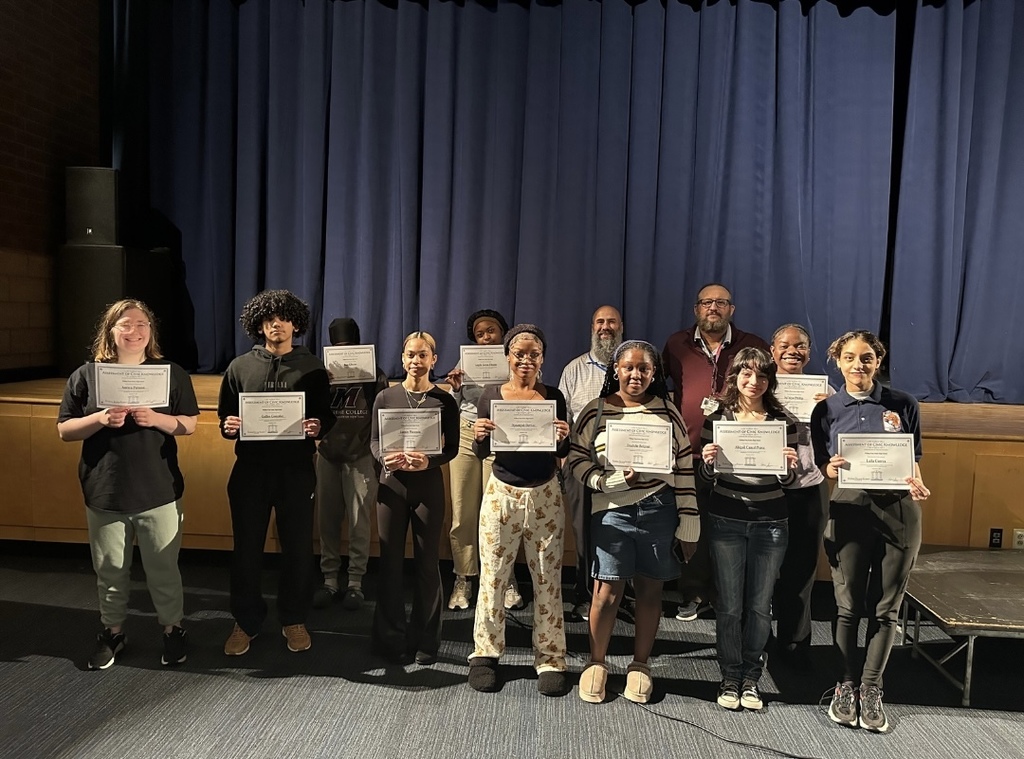 A group of William Penn Senior High School students smiling and holding up certificates with district leadership standing behind them in a school auditorium.