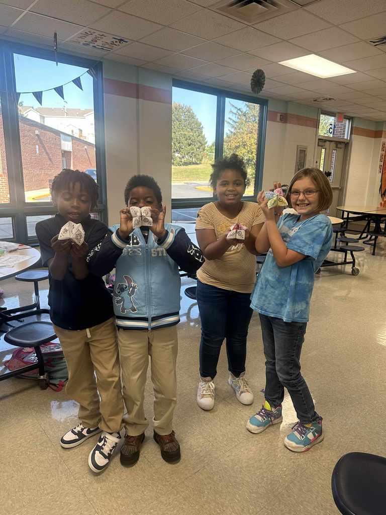 Four students standing in a school cafeteria, each of them are holding a paper fortune teller. 
