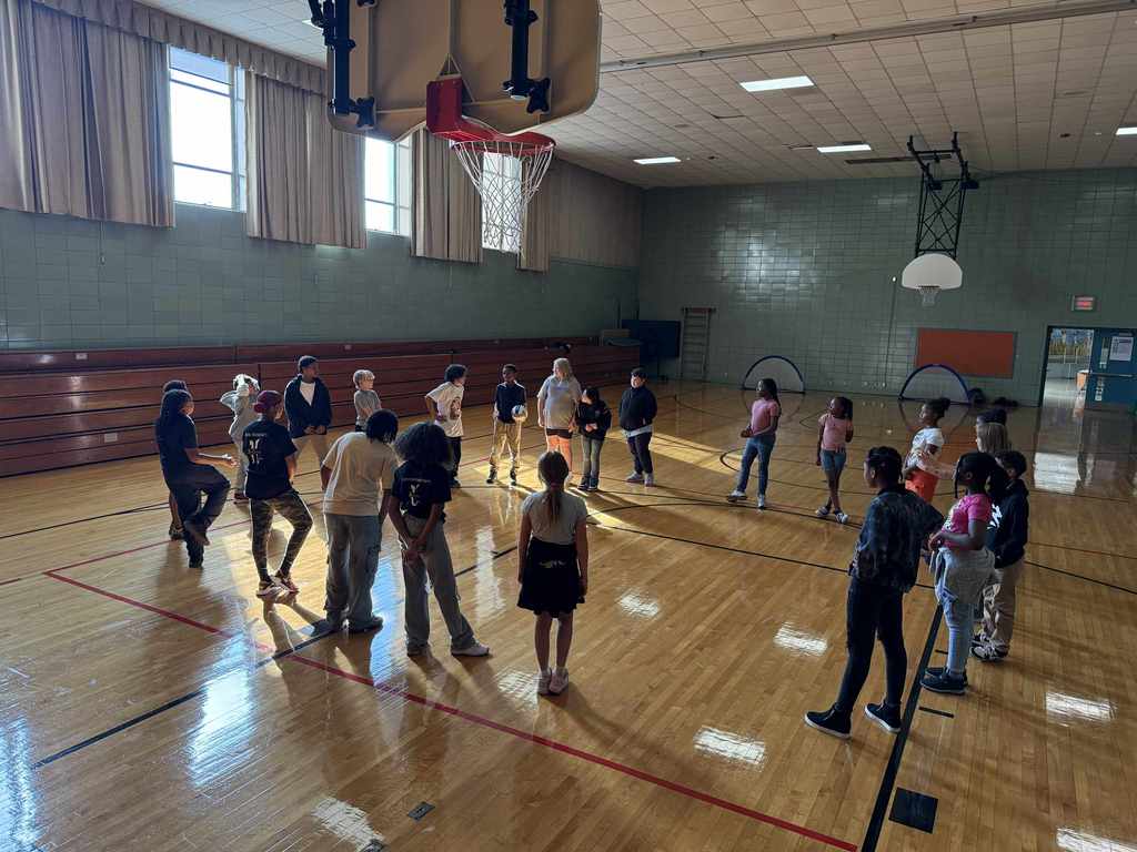 A large group of students standing in a circle in a sunlit gymnasium, underneath a basketball hoop.