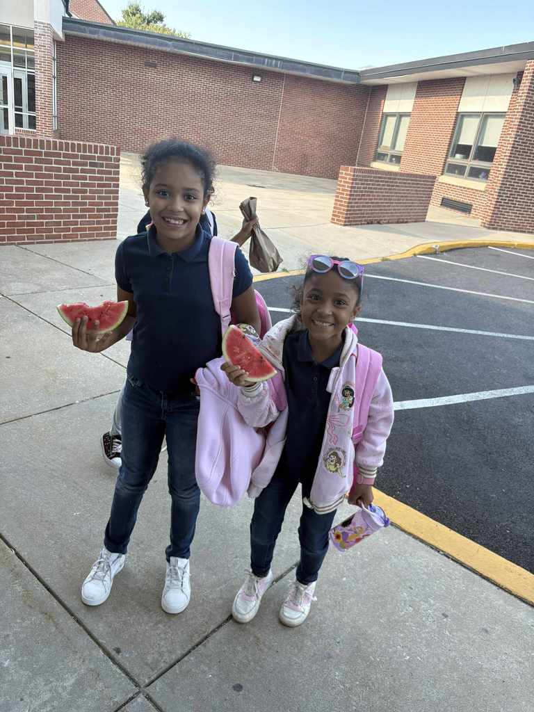 Two students holding up a piece of watermelon while standing outside near a parking lot.