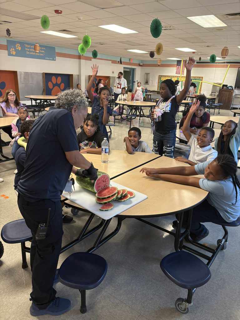 A district staff member is slicing watermelon on a table surrounded by a group of students in a school cafeteria. 