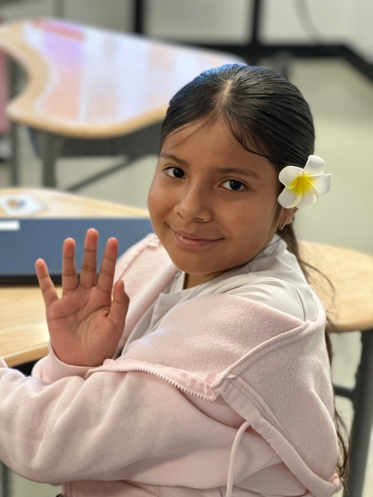 A student wearing a flower in her hand is smiling and waving at a camera while sitting at a desk in a school classroom.