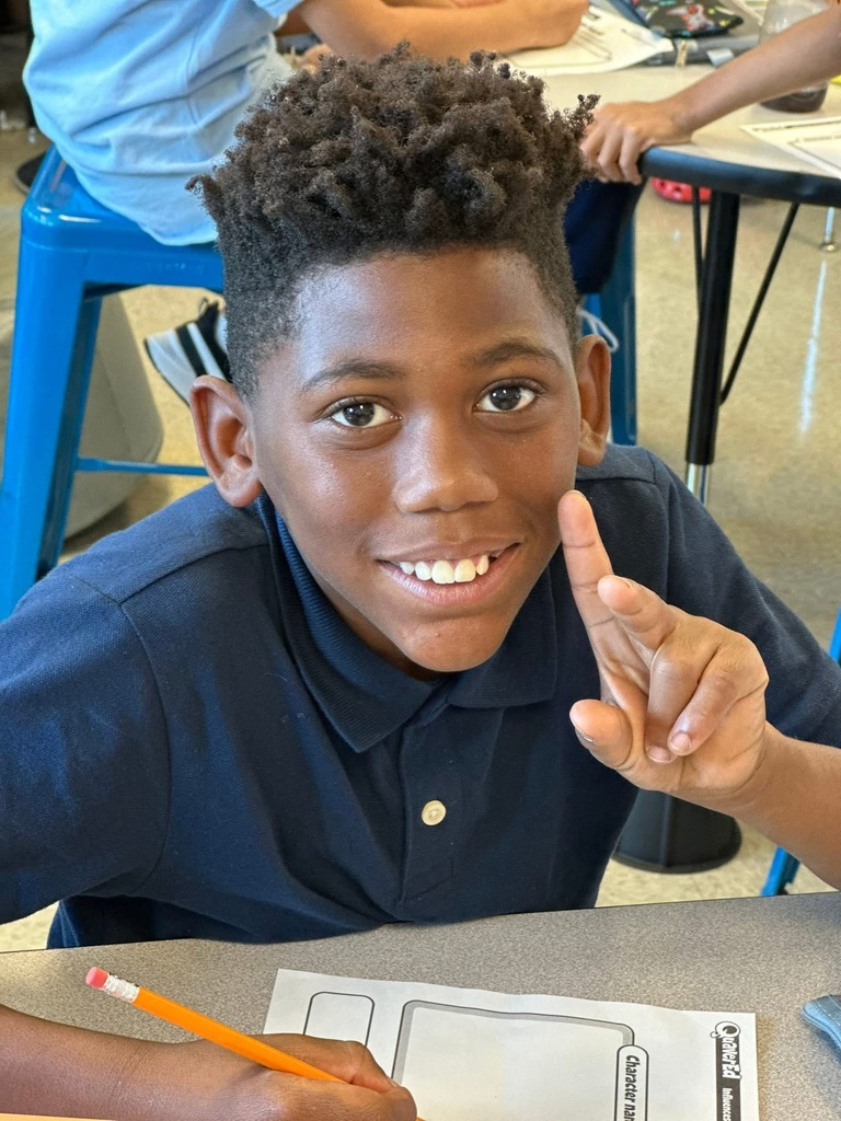 A student smiling and holding up two of his fingers. He has a pencil in the other hand. Other students can be seen in the background.