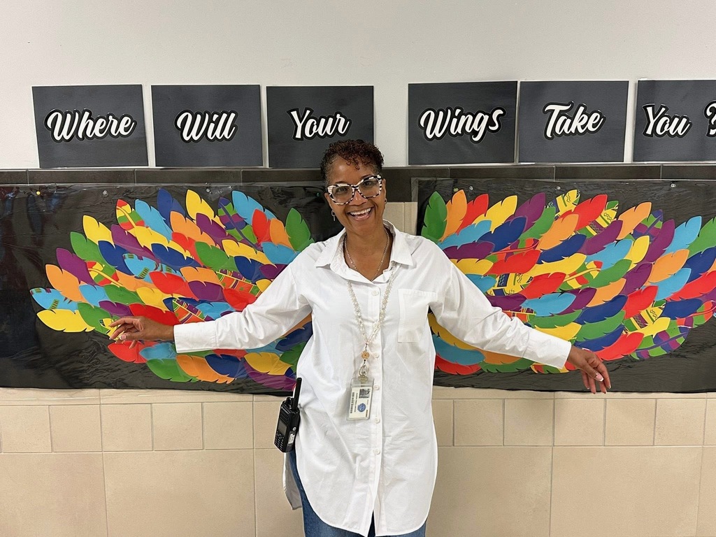 Jackson PreK-8's Principal, Ms. Smallwood smiling and holding her arms up in front of a painting of colorful wings with a text above that says "Where Will Your Wings Take You?"