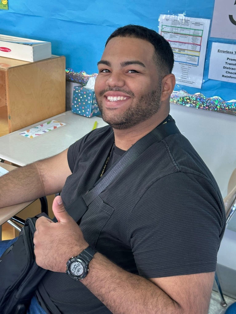 A district staff member smiling and holding his left thumb up while sitting at a desk in a school classroom. 