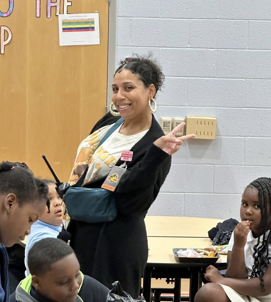 A district staff member smiling and holding up the peace sign while other students are nearby eating food in a school cafeteria.