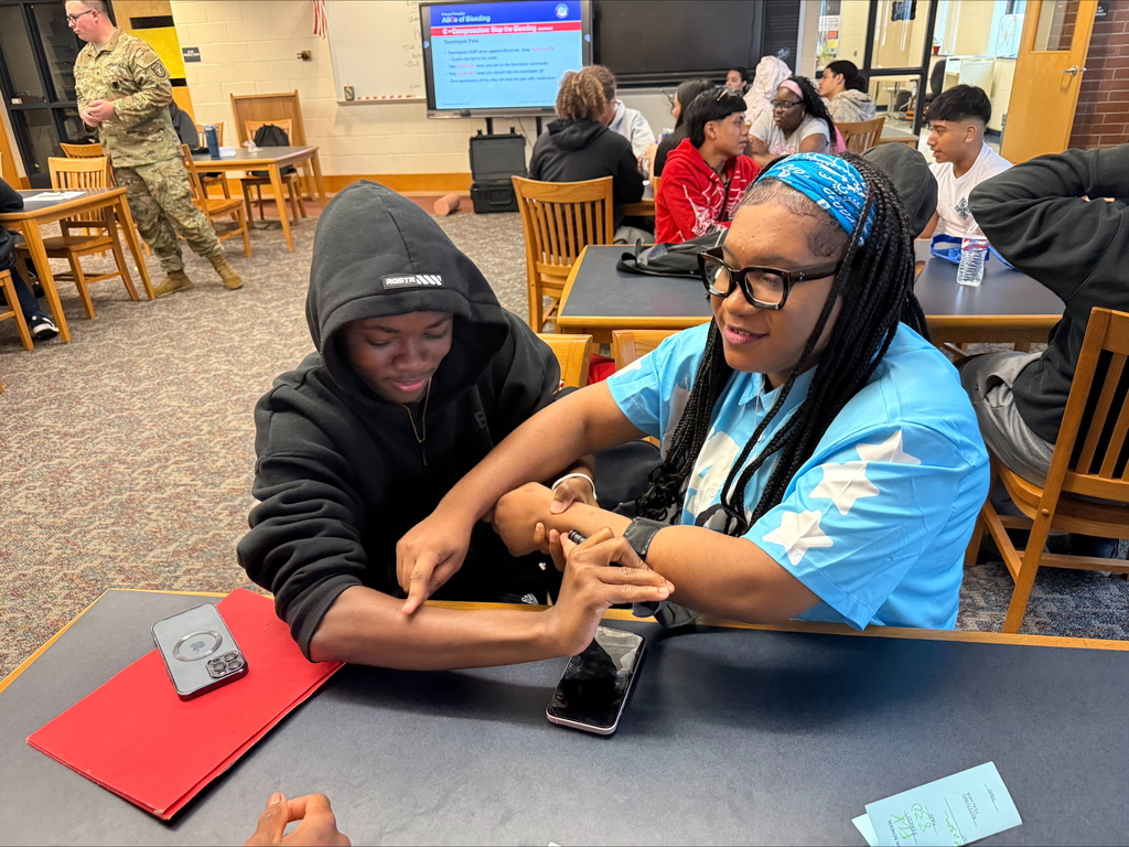 Two students are engaging in a Stop the Bleed Training in a school library. Other people can be seen in the background.