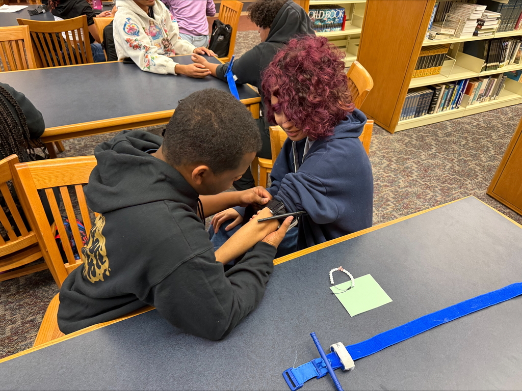 Two students are engaging in a Stop the Bleed Training in a school library. Other people can be seen in the background.