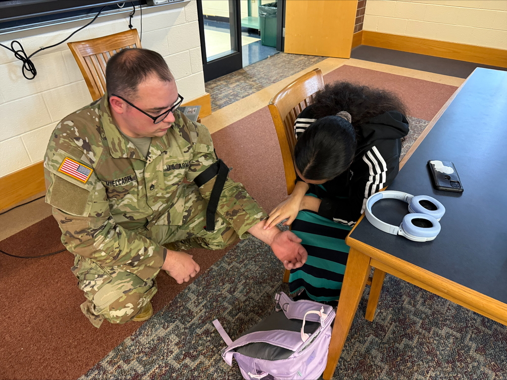 A student and a man wearing military attire are both engaging in a Stop the Bleed Training in a school library. The student is practicing on the military personnel.