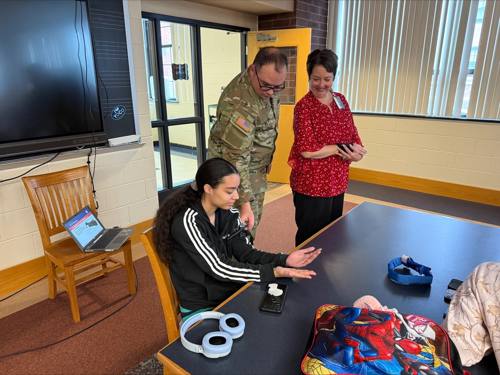 A student is engaging in a Stop the Bleed Training while a district staff member and military personnel are observing her in a school library.