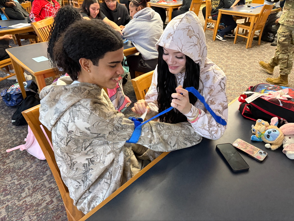Two students are engaging in a Stop the Bleed Training in a school library. Other people can be seen in the background.